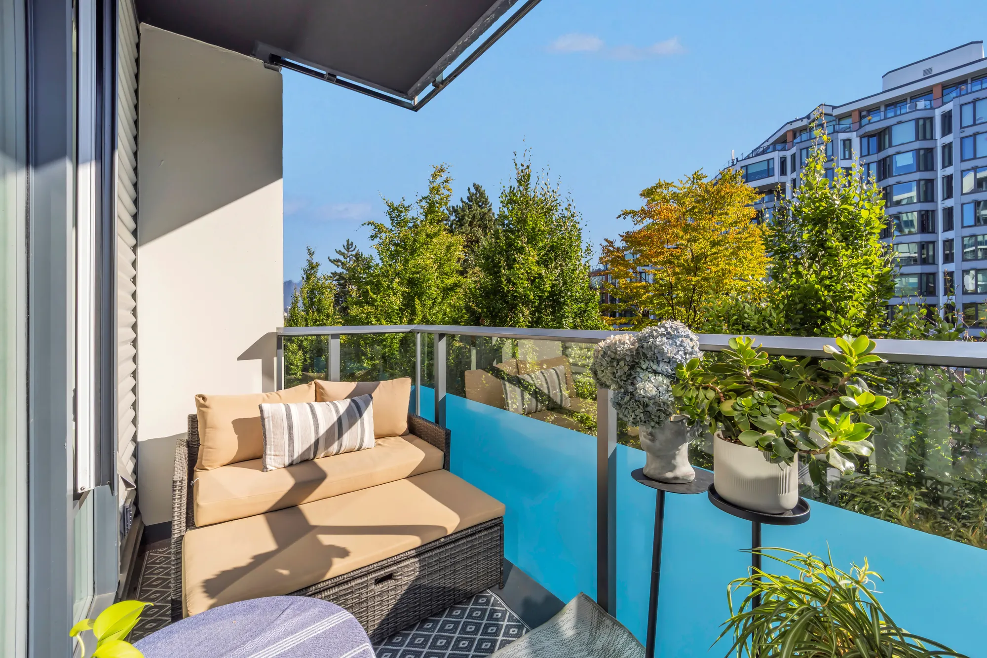 The balcony: a wicker sofa with cushions, planters, a blue glass panel in the railing, the courtyard's trees and the building across it