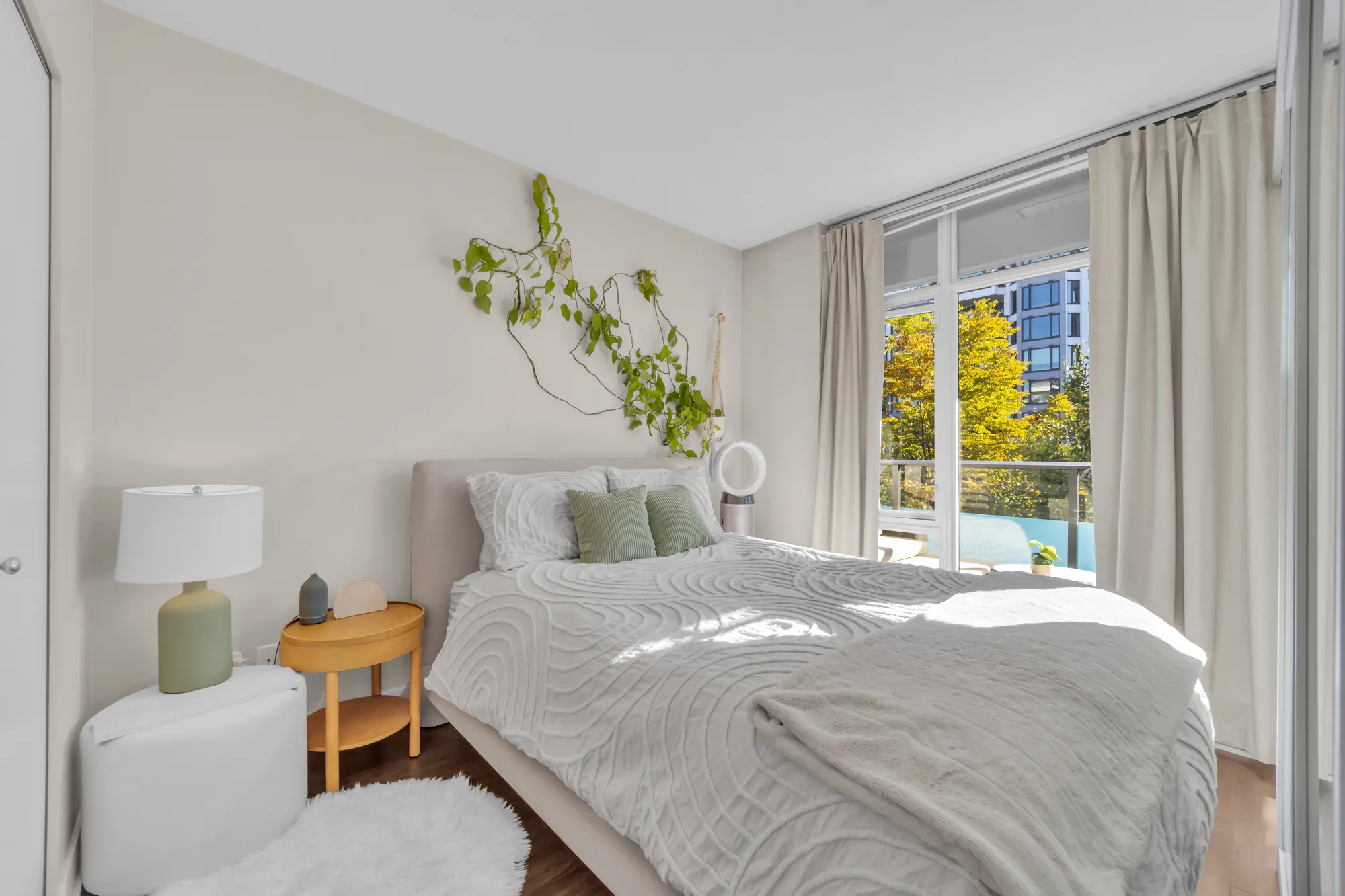 The bedroom: a bed with a white quilted cover by a window with curtains, a lamp on a night table, a plant on the wall, the sliding glass wall on the right