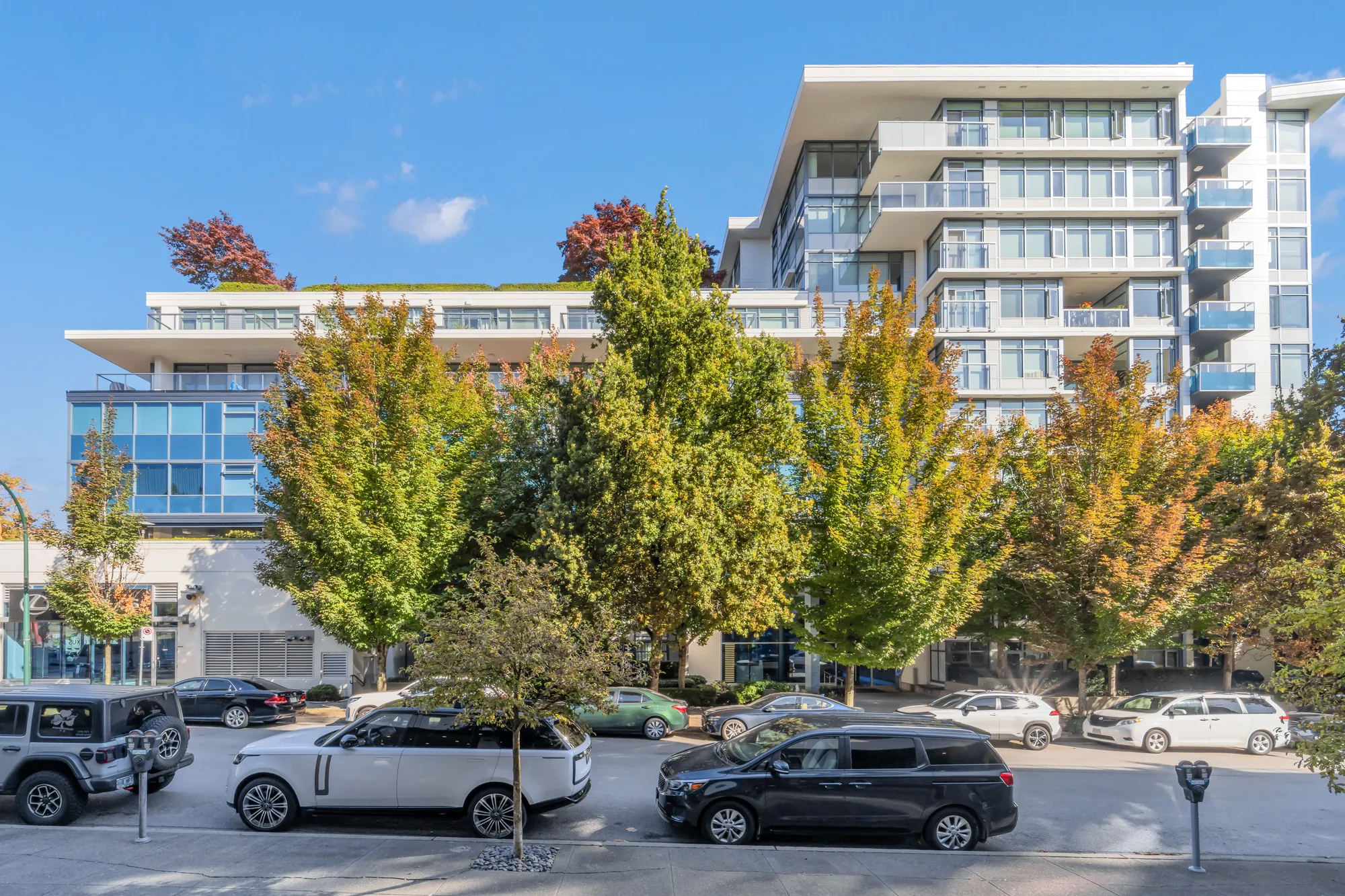 Kits360 from 7th Avenue: the glass and concrete building behind trees in autumn colour, cars parked along the street