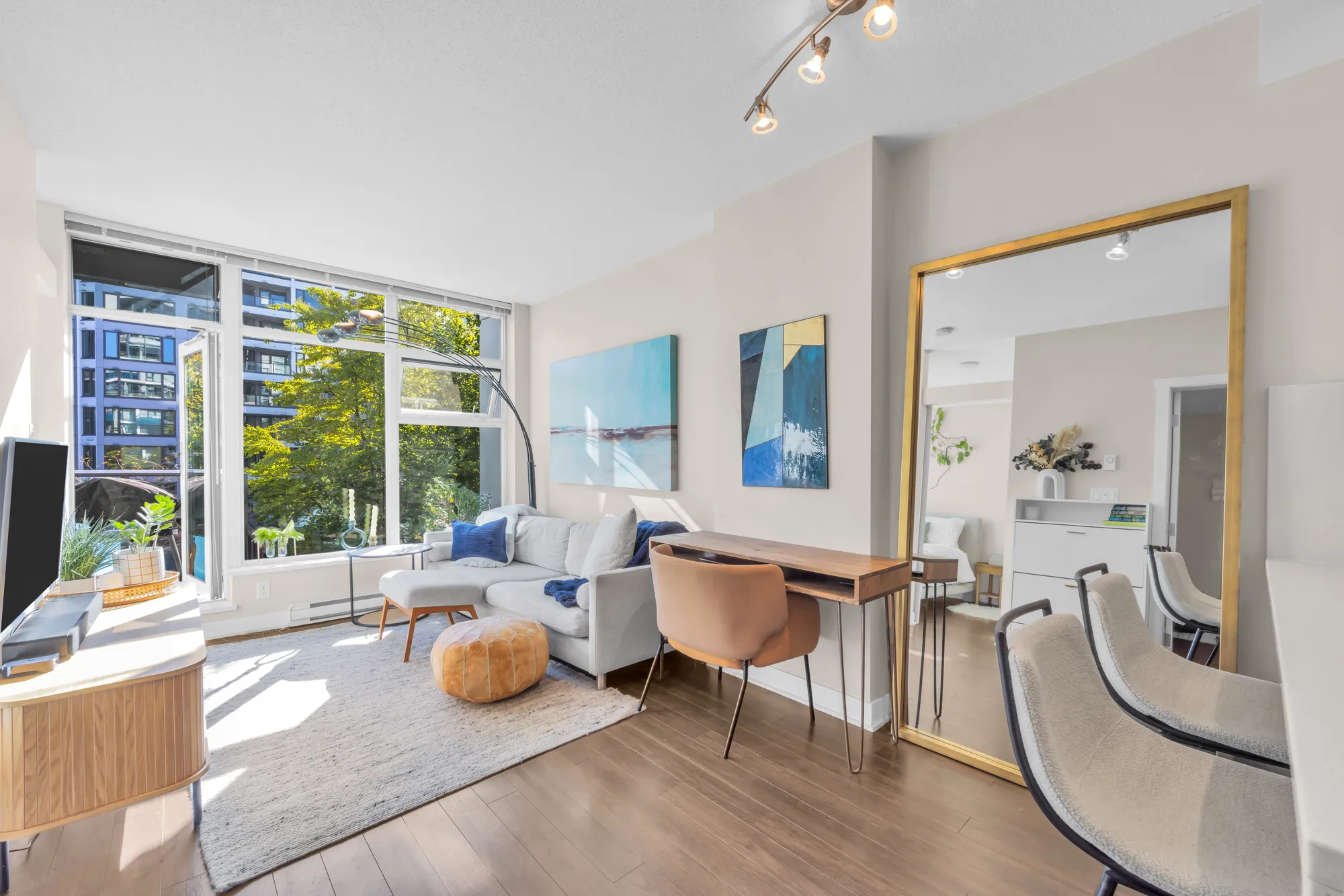 The living room of 317-1777 West 7th Avenue: a white sofa and a tan leather chair under a corner of windows, two blue paintings, the courtyard's trees outside