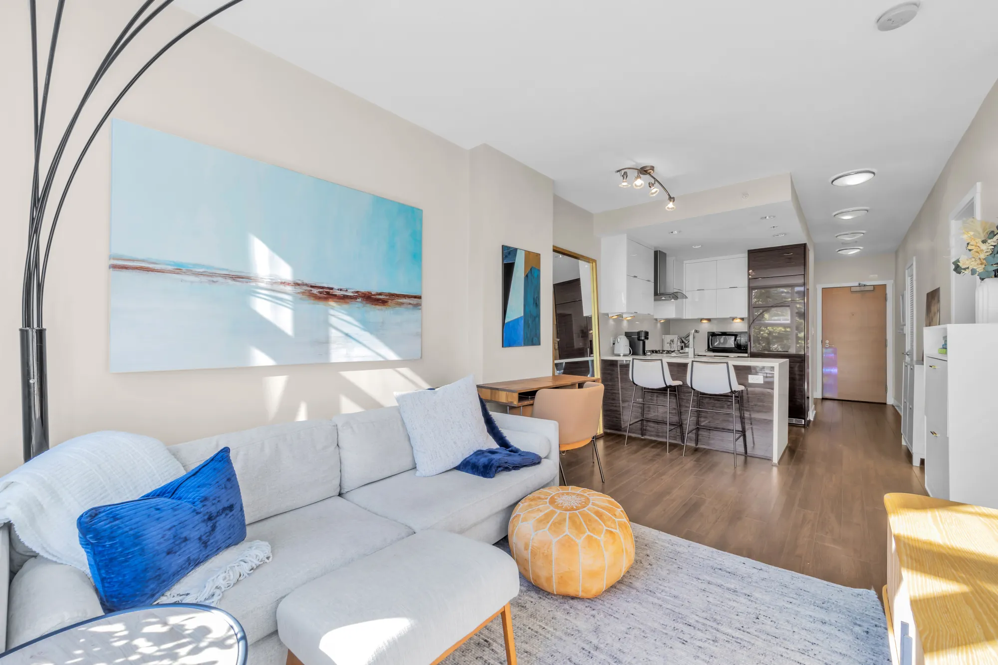 The living and dining rooms toward the kitchen: the white sofa, a pouf, the paintings, the peninsula with stools, the door beyond